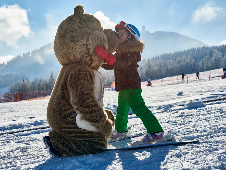 Ein junges Mädchen auf einem Snowboard umarmt das Ar"Bär" Masskotchen des Großen Arbers.
